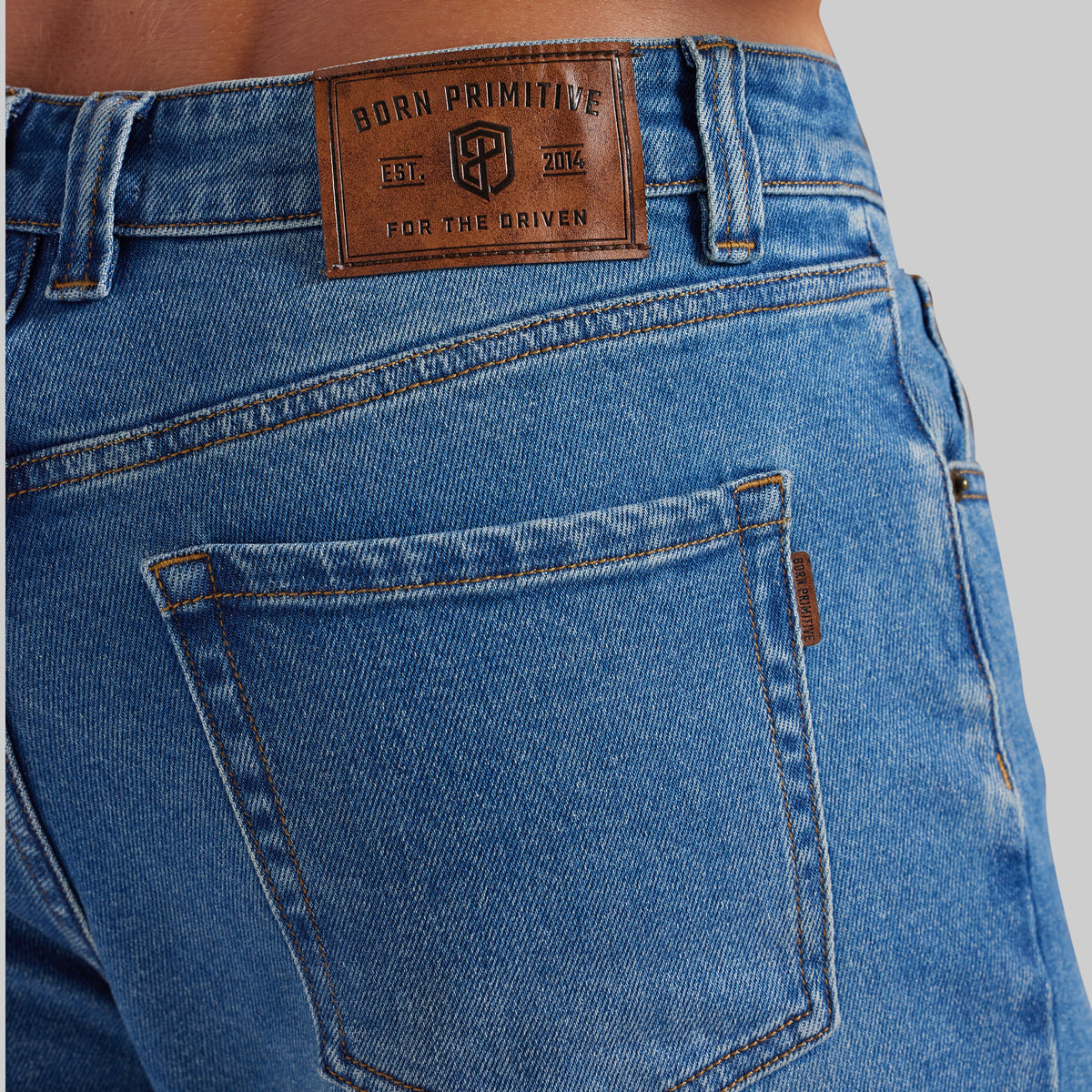 Close-up of the back waistband and pocket area of blue denim jeans on a model, showing a brown leather patch with the Born Primitive logo.