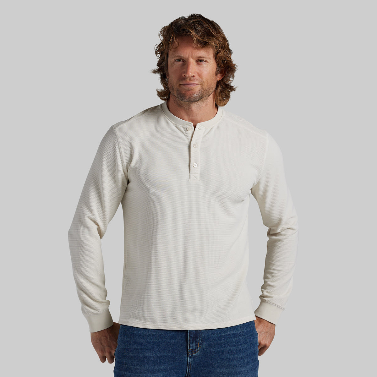 A man wearing a cream-colored long-sleeve henley shirt with blue jeans, standing against a solid gray studio background.
