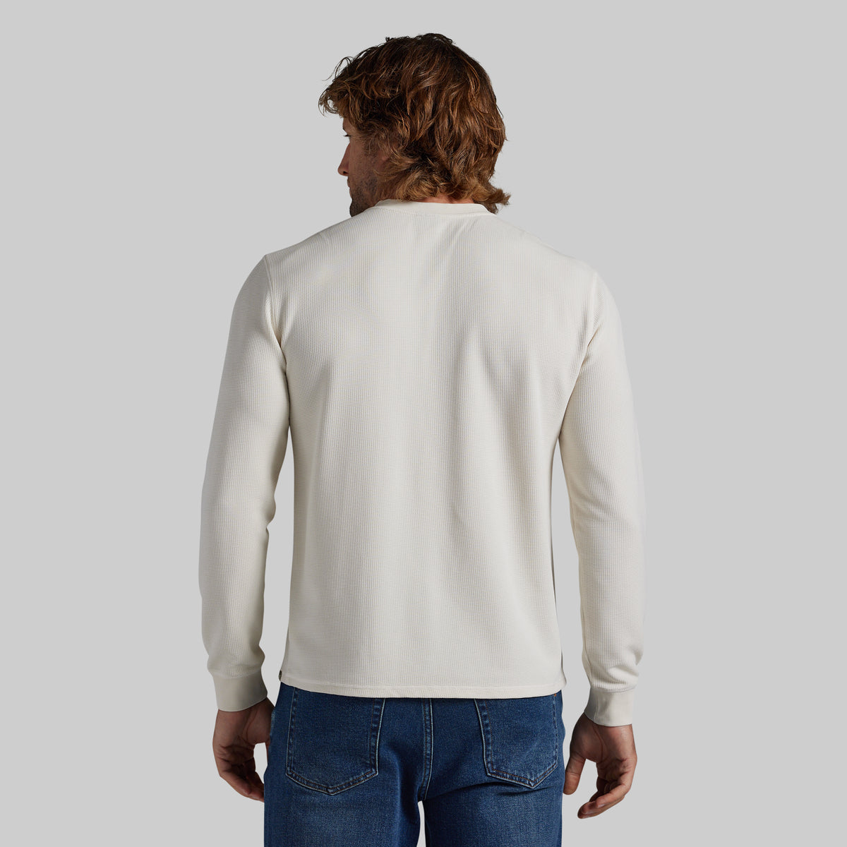 A man viewed from behind wearing a plain off-white long-sleeve crewneck sweater and blue jeans, set against a solid light gray background.