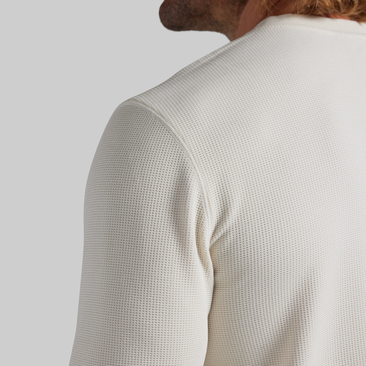Close-up back view of a person wearing a textured off-white waffle-knit long-sleeve top against a light gray studio background.