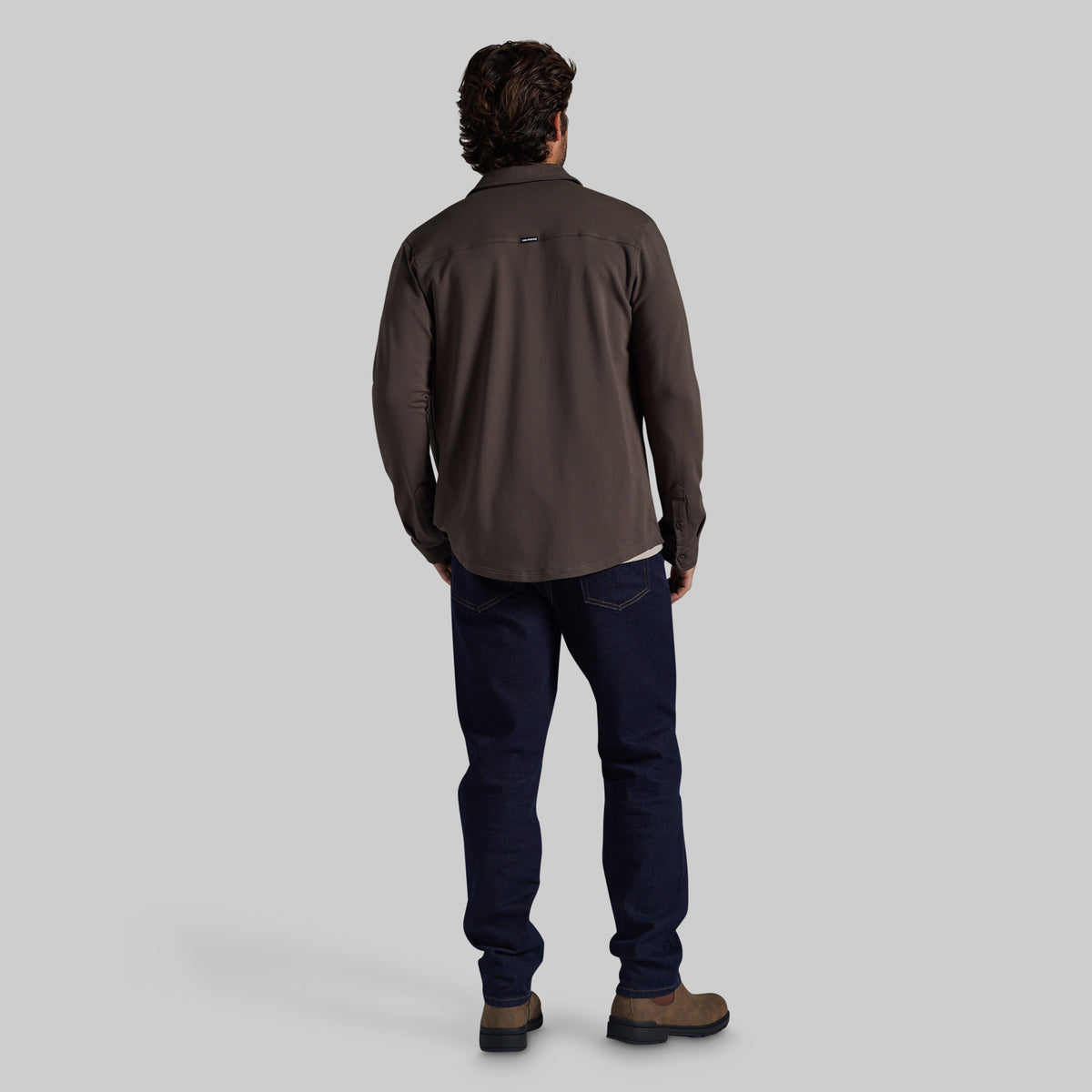 A man viewed from behind wearing a dark brown long-sleeve performance shirt, navy blue jeans, and brown hiking boots against a solid gray studio background.
