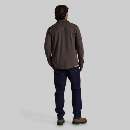 A man viewed from behind wearing a dark brown long-sleeve performance shirt, navy blue jeans, and brown hiking boots against a solid gray studio background.