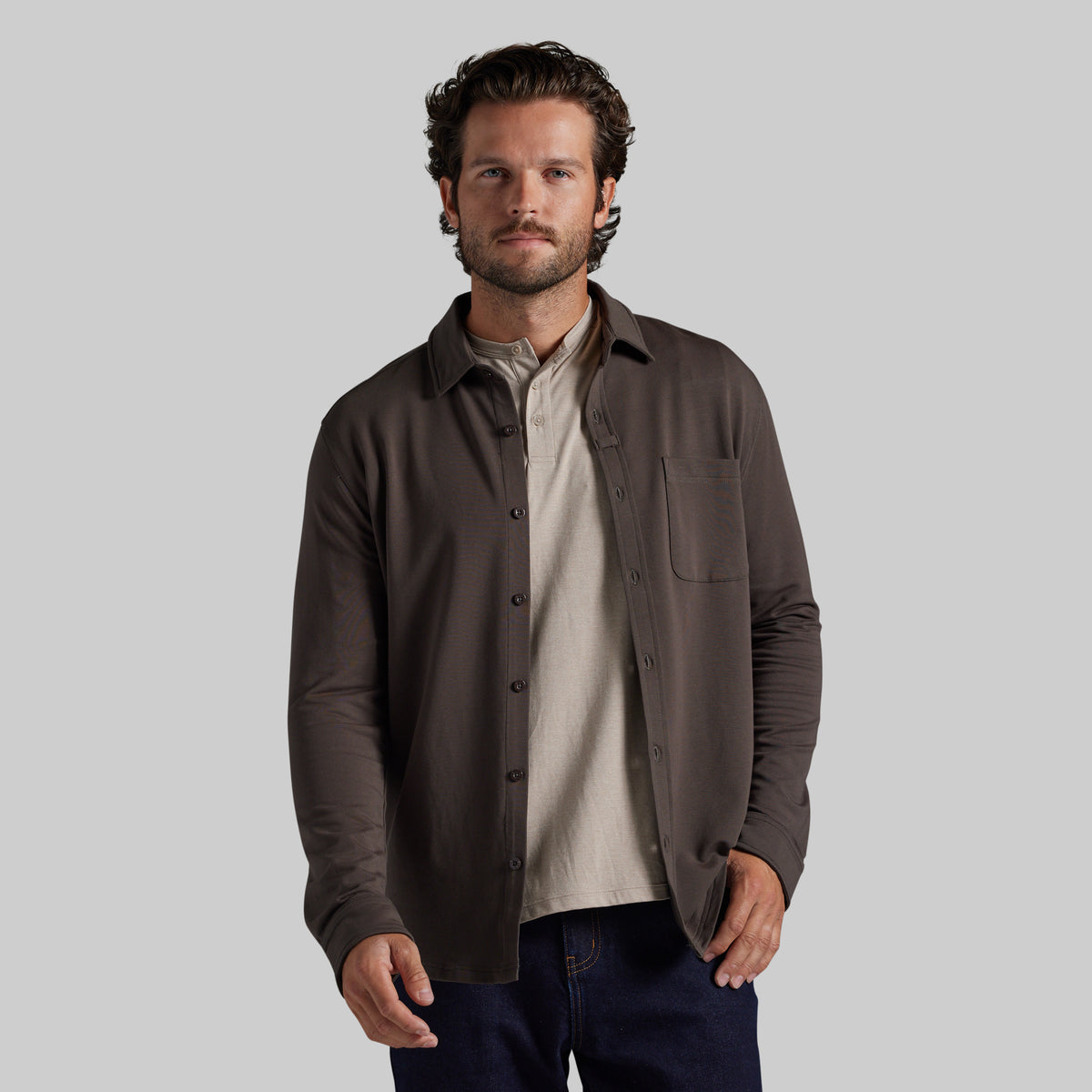 A man wearing a dark brown long-sleeve button-up overshirt over a light beige shirt and dark blue jeans, photographed against a solid light gray background.
