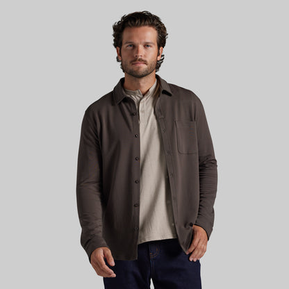 A man wearing a dark brown long-sleeve button-up overshirt over a light beige shirt and dark blue jeans, photographed against a solid light gray background.