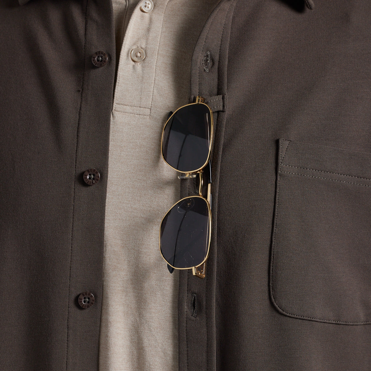 Close-up of a dark olive brown overshirt worn over a light beige polo shirt, with gold-framed sunglasses hanging from the chest pocket.