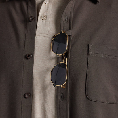 Close-up of a dark olive brown overshirt worn over a light beige polo shirt, with gold-framed sunglasses hanging from the chest pocket.