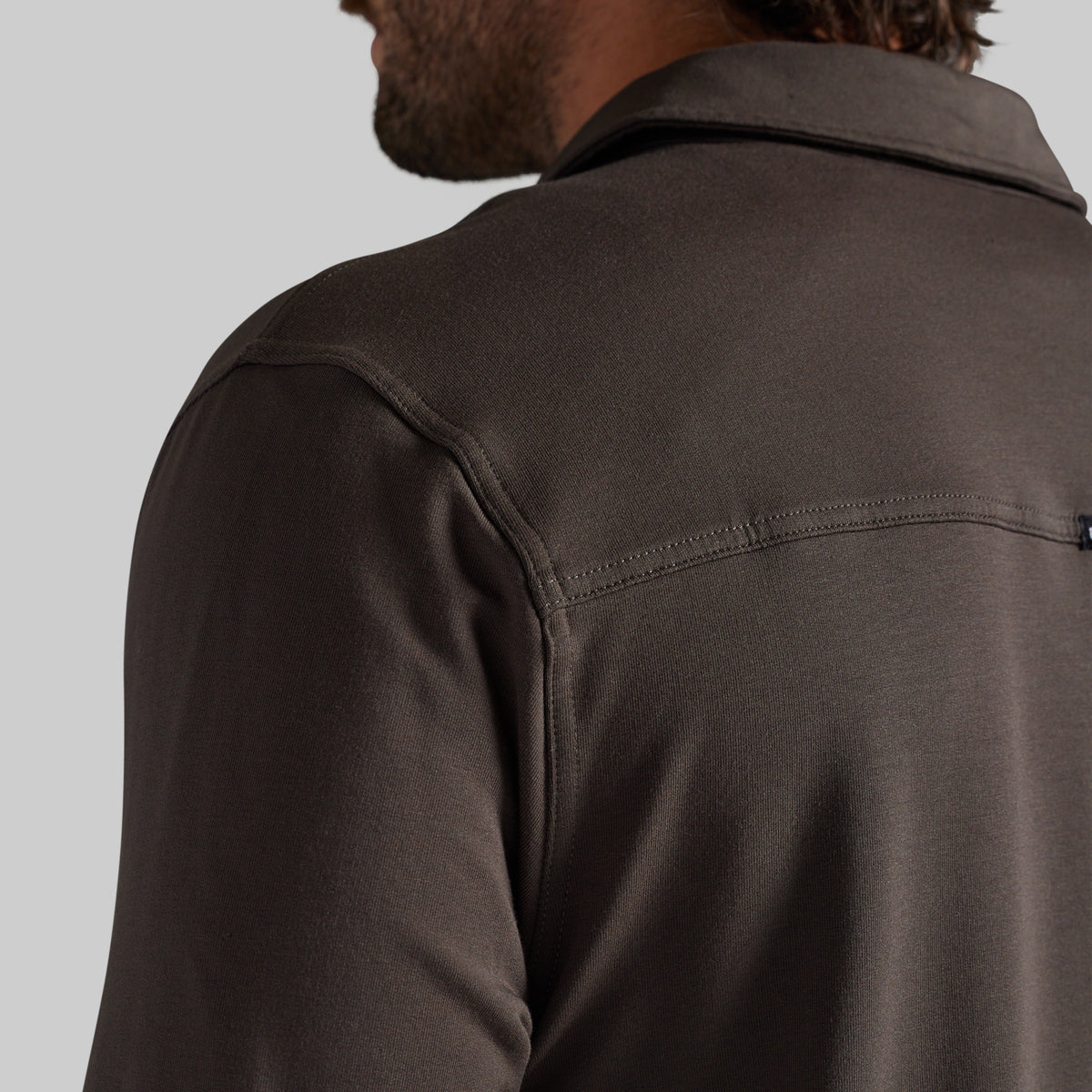 Close-up back view of a man wearing a dark brown long-sleeve collared shirt against a light gray background.