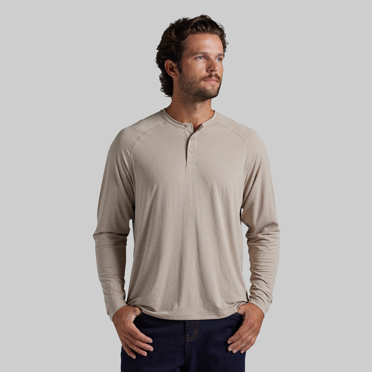 A man wearing a light beige long-sleeve henley shirt with a buttoned collar, paired with dark blue jeans, standing against a solid gray background.