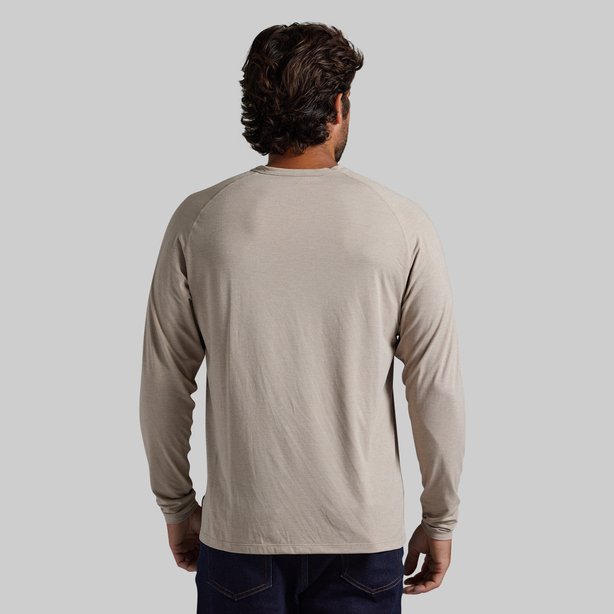 A man viewed from behind wearing a light beige long-sleeve raglan t-shirt and dark blue jeans, set against a solid light gray background.