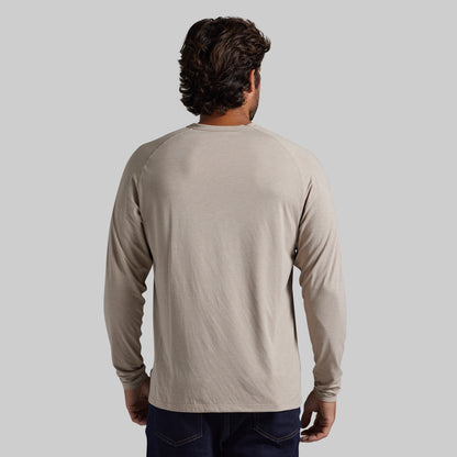 A man viewed from behind wearing a light beige long-sleeve raglan t-shirt and dark blue jeans, set against a solid light gray background.