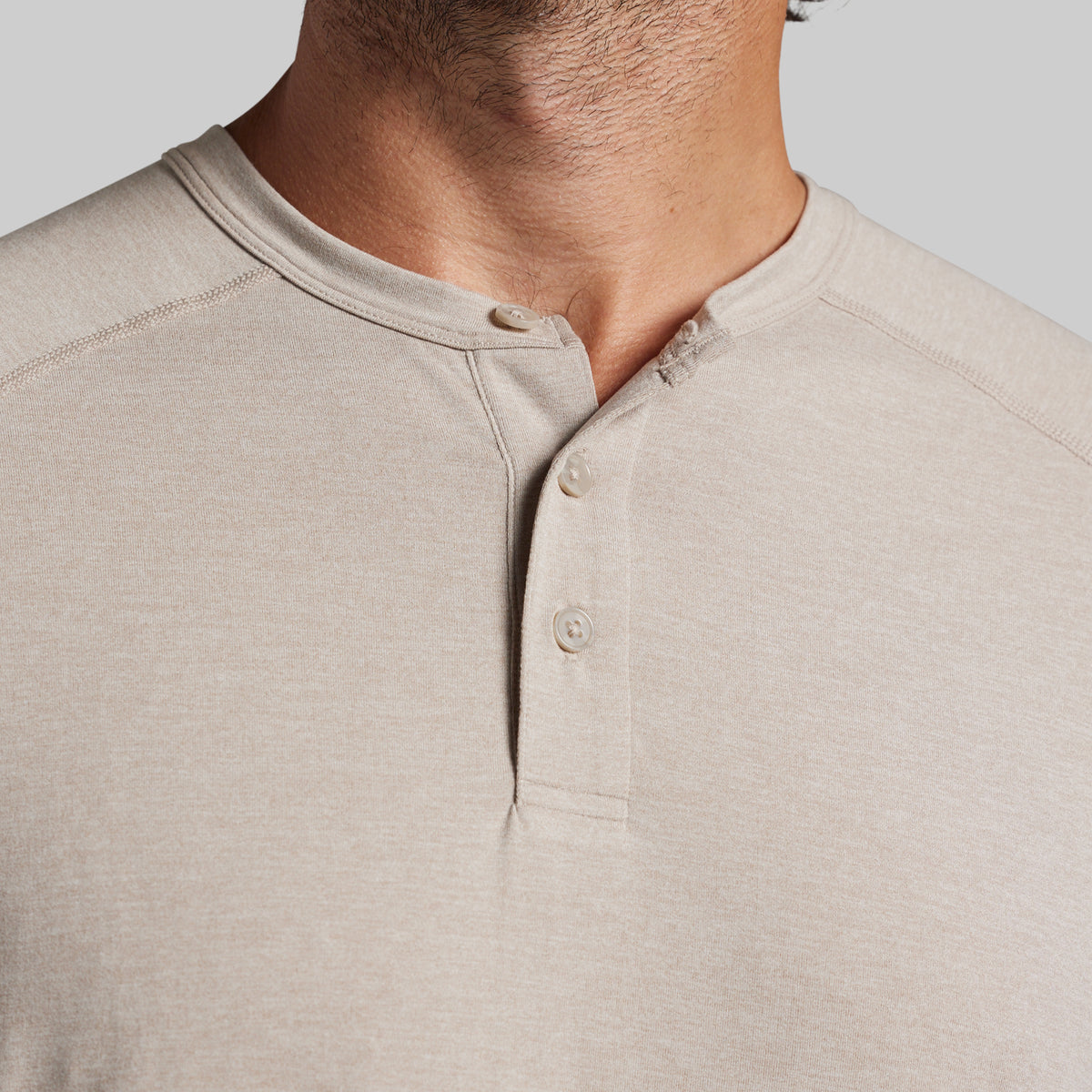 Close-up of a man wearing a light beige heather henley shirt, showing the collar, three-button placket, and chest fabric against a plain light gray background.