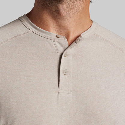 Close-up of a man wearing a light beige heather henley shirt, showing the collar, three-button placket, and chest fabric against a plain light gray background.