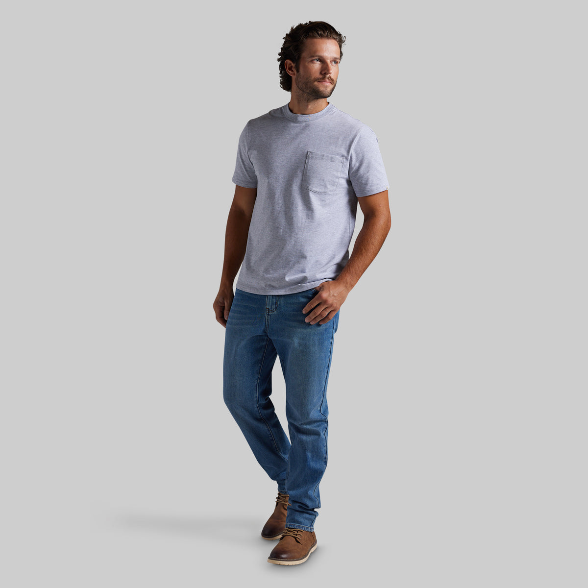 A man wearing a light gray short-sleeve crewneck t-shirt with a chest pocket, blue jeans, and brown shoes, standing against a solid gray background.