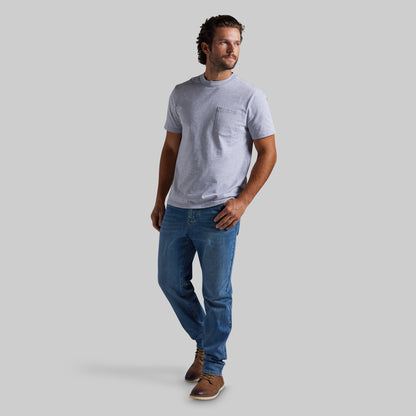 A man wearing a light gray short-sleeve crewneck t-shirt with a chest pocket, blue jeans, and brown shoes, standing against a solid gray background.