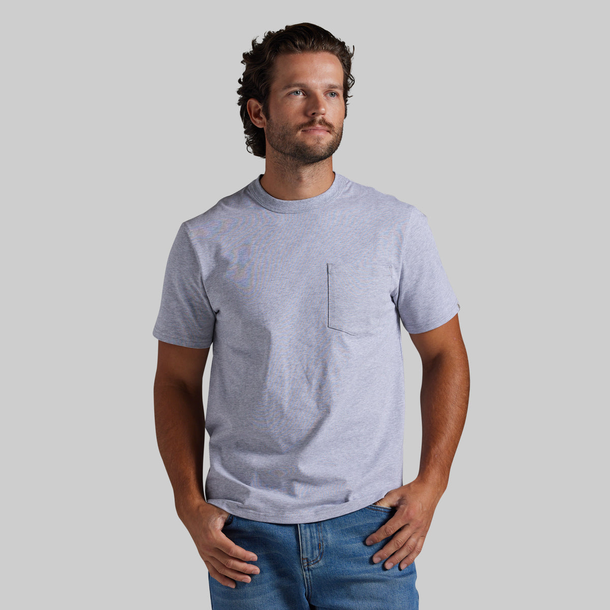 A man wearing a light heather gray short-sleeve crewneck t-shirt with a chest pocket, paired with blue jeans, standing against a solid gray background.