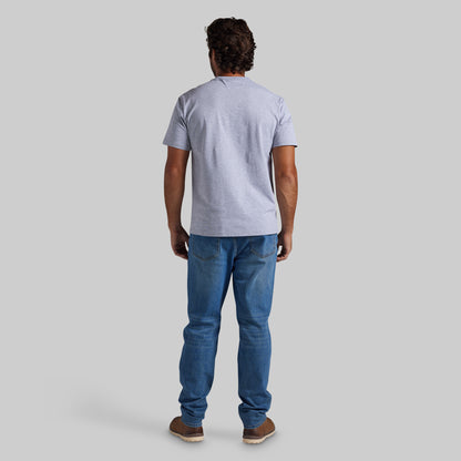 A man viewed from behind wearing a light heather gray t-shirt, blue jeans, and brown shoes, standing against a solid gray studio background.