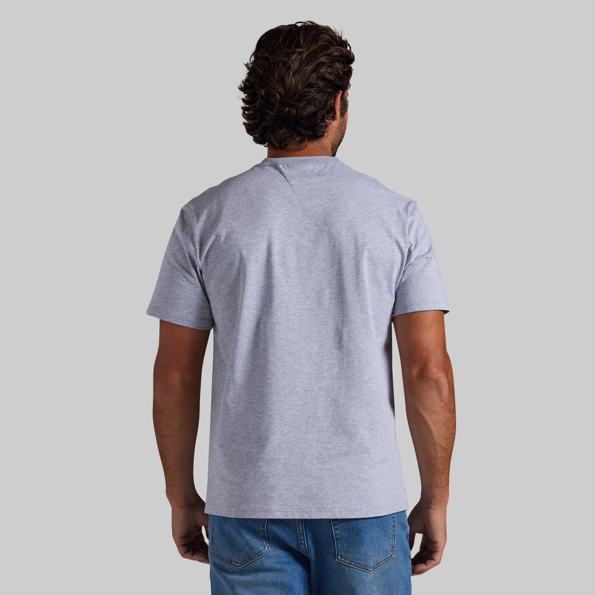 A man viewed from behind wearing a light heather gray short-sleeve t-shirt and blue jeans, standing against a solid light gray background.