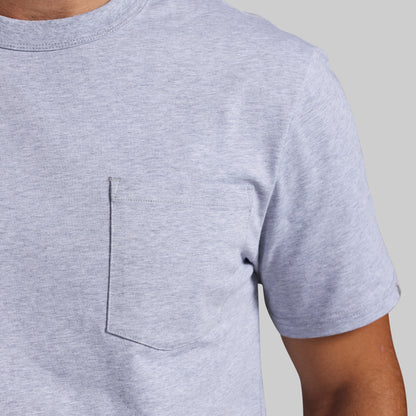 Close-up of a person wearing a light heather gray short-sleeve crewneck t-shirt with a chest pocket, set against a solid light gray background.