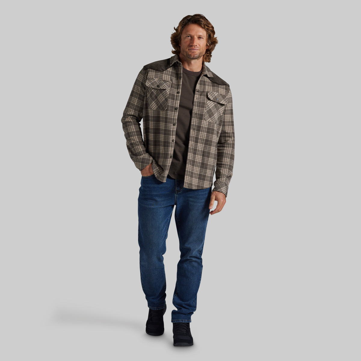 A man modeling a brown and beige plaid western-style flannel shirt jacket over a dark t-shirt and blue jeans, standing against a solid gray studio background.