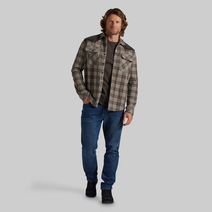 A man modeling a brown and beige plaid western-style flannel shirt jacket over a dark t-shirt and blue jeans, standing against a solid gray studio background.