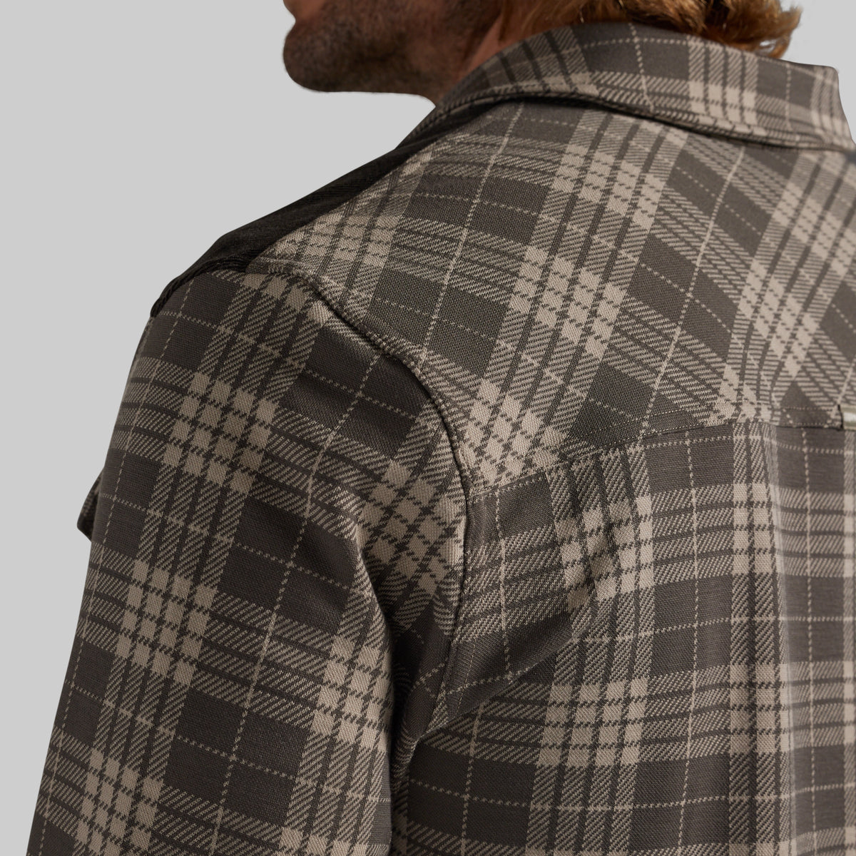 Close-up of a man wearing a brown and beige plaid overshirt, showing the collar, shoulder, and fabric texture against a solid gray background.
