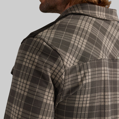 Close-up of a man wearing a brown and beige plaid overshirt, showing the collar, shoulder, and fabric texture against a solid gray background.