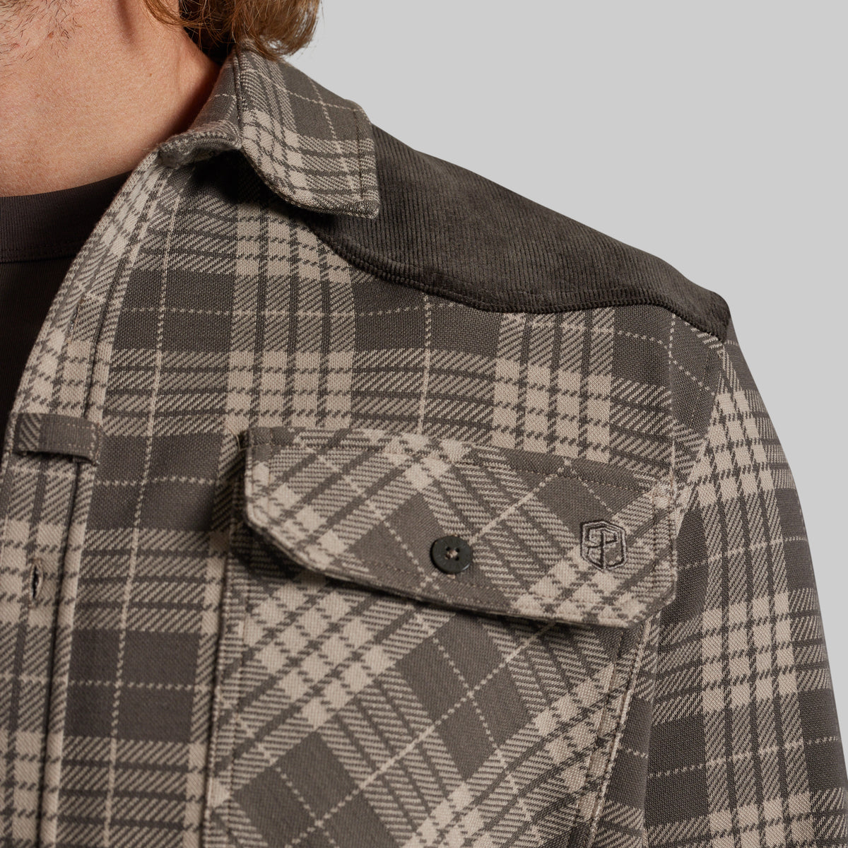 Close-up of a person wearing a plaid overshirt with a dark corduroy yoke and a flap chest pocket, against a light gray background.