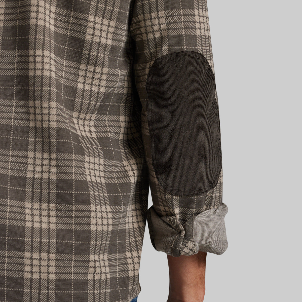 Close-up of a plaid flannel shirt with a dark corduroy elbow patch and rolled-up sleeve, shown on a model against a light gray background.