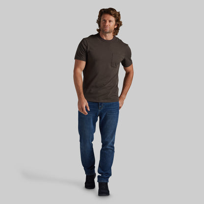 A man modeling a dark brown short-sleeve crewneck pocket t-shirt with blue jeans and black shoes, standing against a solid gray background.