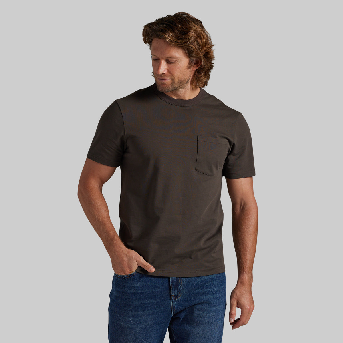 A man wearing a dark brown short-sleeve crewneck pocket t-shirt and blue jeans, standing against a solid gray background.