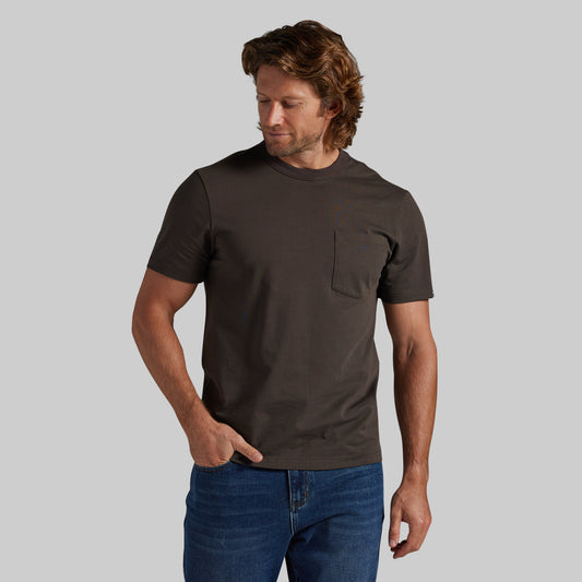 A man wearing a dark brown short-sleeve crewneck pocket t-shirt and blue jeans, standing against a solid gray background.