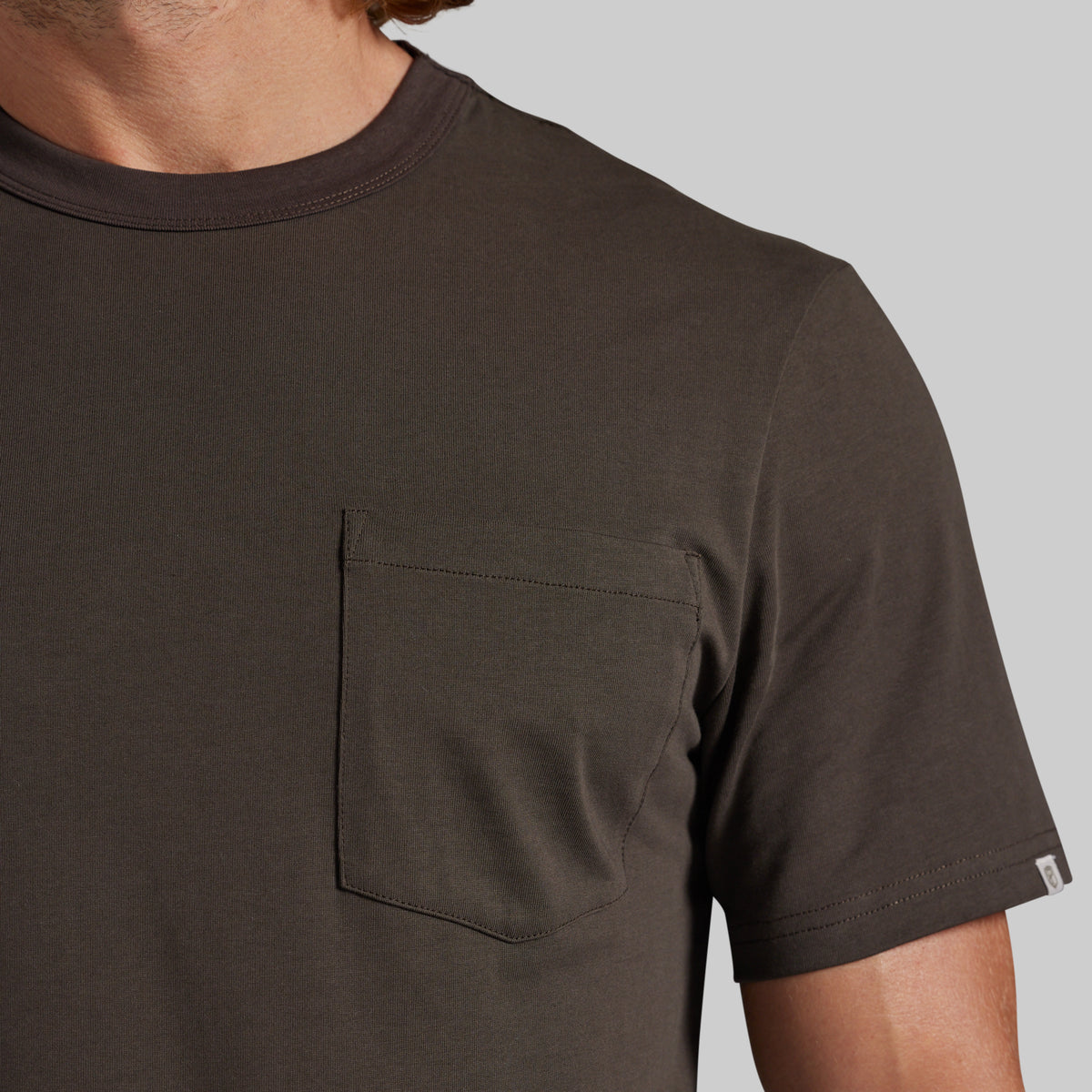 Close-up of a man wearing a dark brown short-sleeve crewneck t-shirt with a chest pocket, set against a solid gray background.