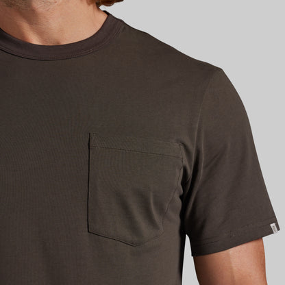 Close-up of a man wearing a dark brown short-sleeve crewneck t-shirt with a chest pocket, set against a solid gray background.
