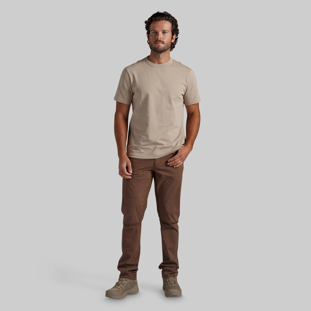 A man modeling a beige crew-neck t-shirt and brown pants against a solid gray background.