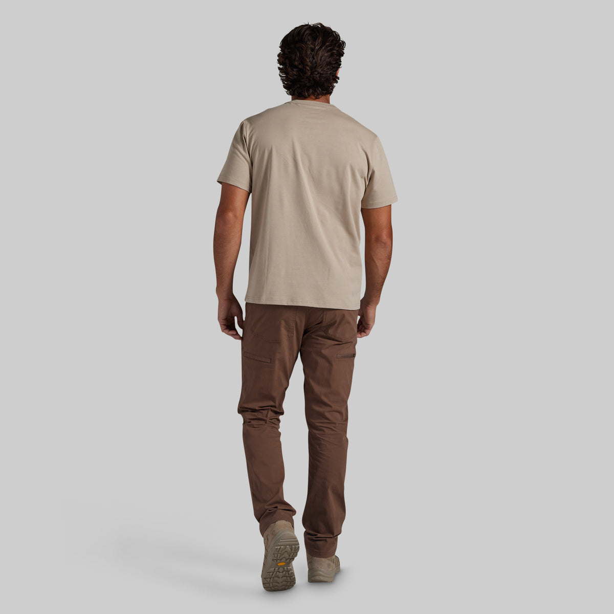 A man shown from behind wearing a beige short-sleeve t-shirt, brown pants, and beige shoes, walking against a solid light gray background.