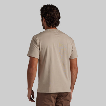 A man viewed from behind wearing a light beige short-sleeve t-shirt, paired with brown pants, against a solid gray background.