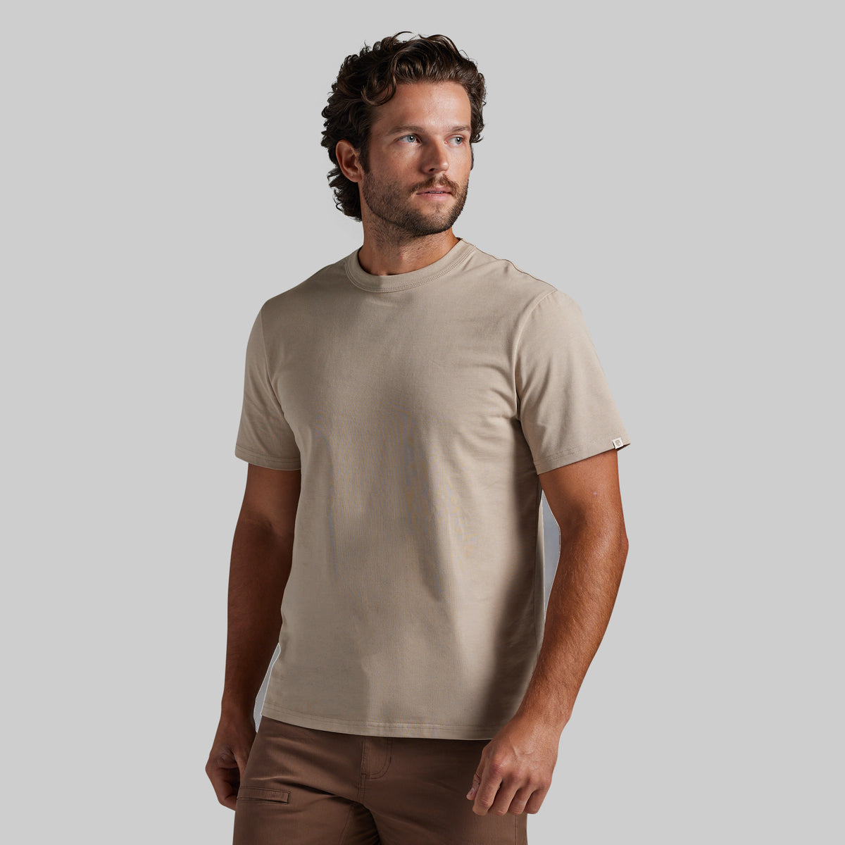 A man wearing a light beige crew-neck t-shirt and brown pants, standing against a solid gray studio background.