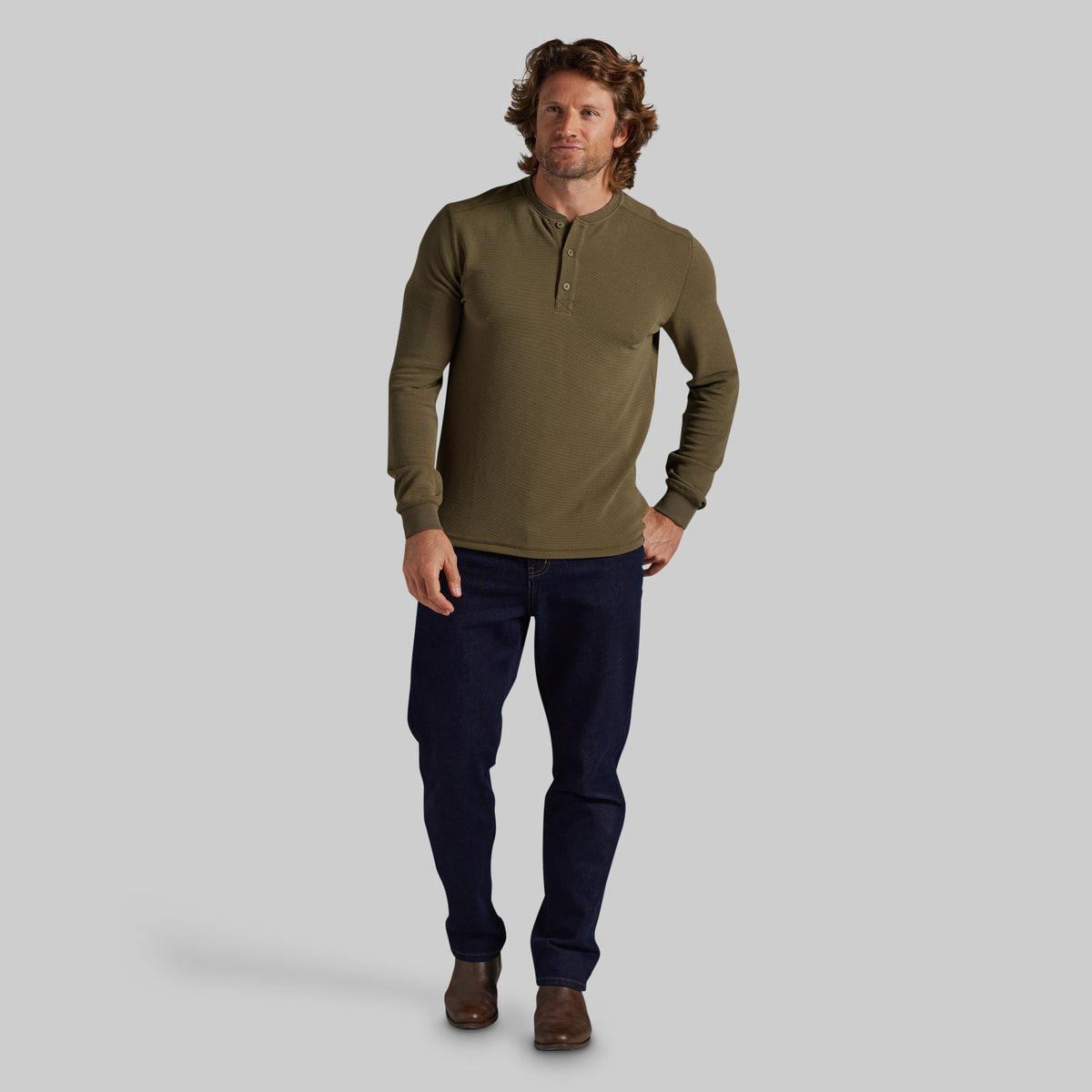 A man modeling a long-sleeve olive green henley shirt with dark navy pants and brown shoes, standing against a solid gray studio background.