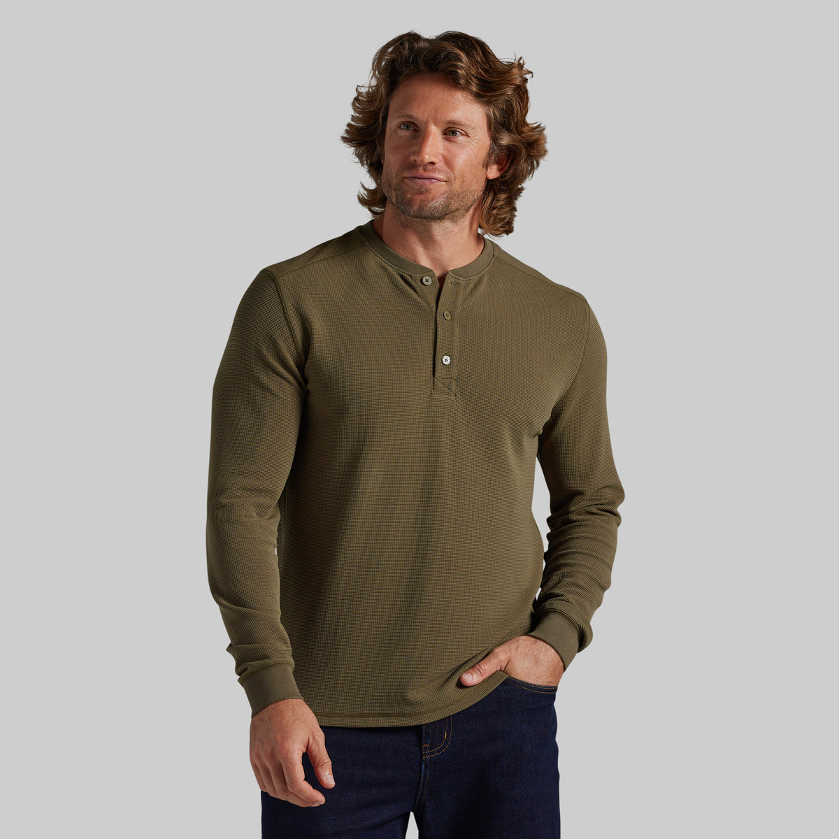 A man wearing a long-sleeve henley shirt in olive green, paired with dark blue jeans, standing against a solid gray background.