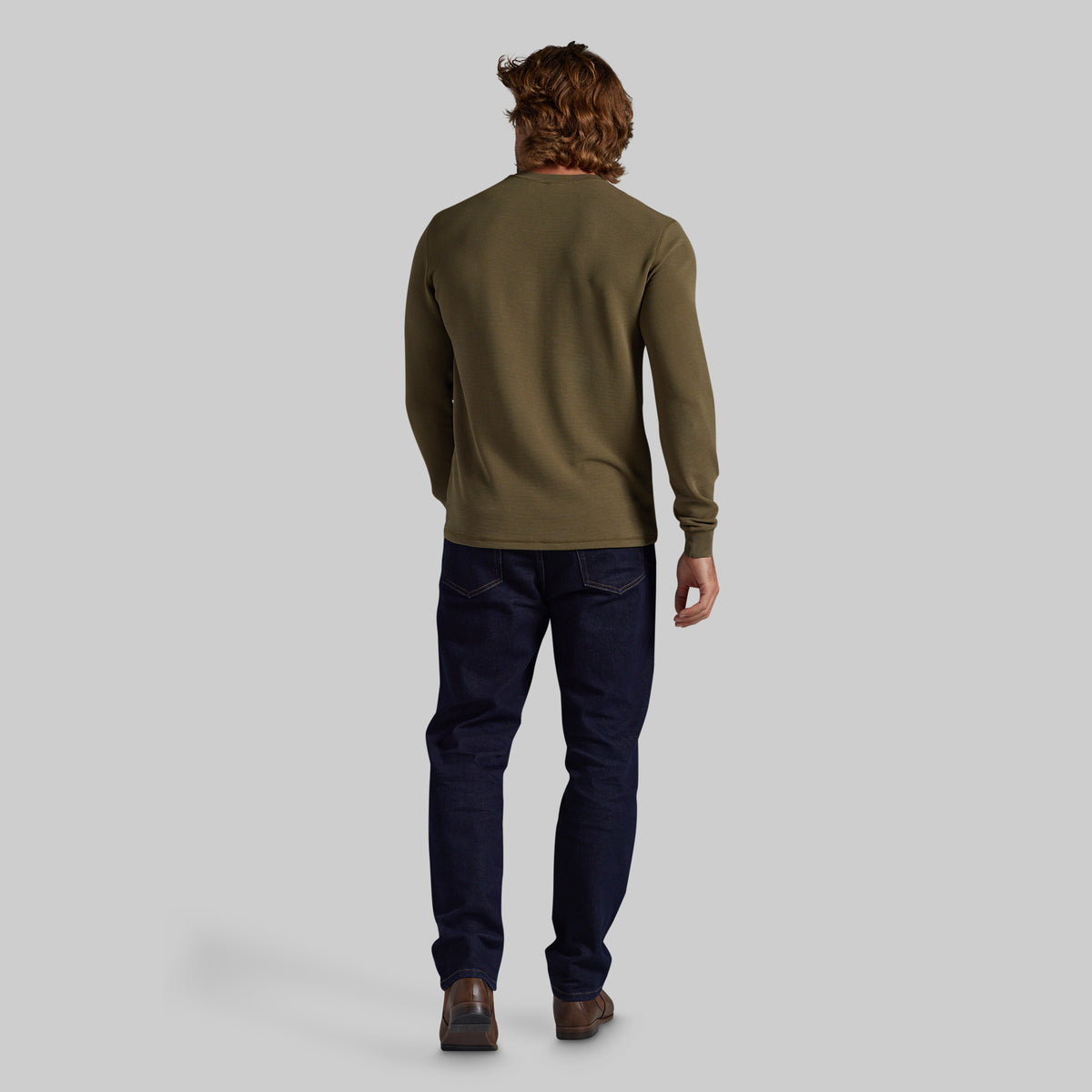 A man viewed from behind wearing an olive green long-sleeve crewneck shirt, dark navy blue jeans, and brown leather shoes, standing against a solid light gray background.