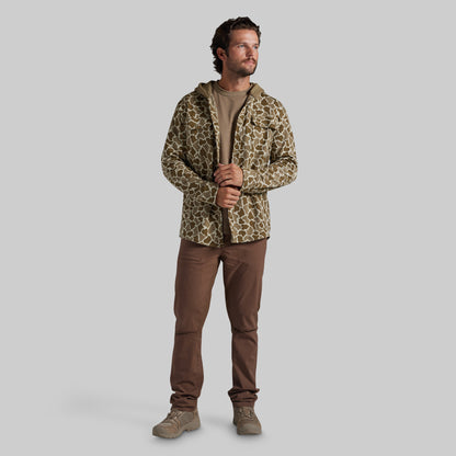 A man modeling a hooded flannel with a brown and beige animal print pattern, worn over a brown t-shirt and brown pants, against a solid gray background.