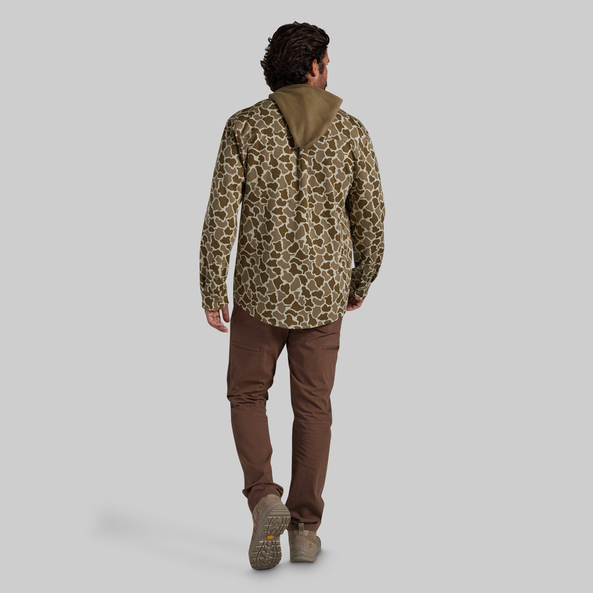 A man shown from behind wearing a hooded flannel with a brown and tan animal-style pattern, brown pants, and tan boots, standing against a light gray studio background.