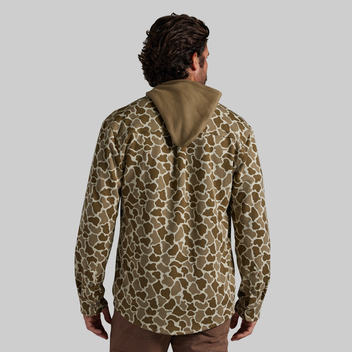 A man viewed from behind wearing a long-sleeve hooded flannel with a frog camo brown and beige pattern, paired with brown pants, against a solid gray studio background.