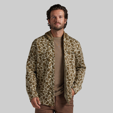 A man wearing a brown and beige frog camo hooded shirt over a tan shirt and brown pants, standing against a solid gray studio background.