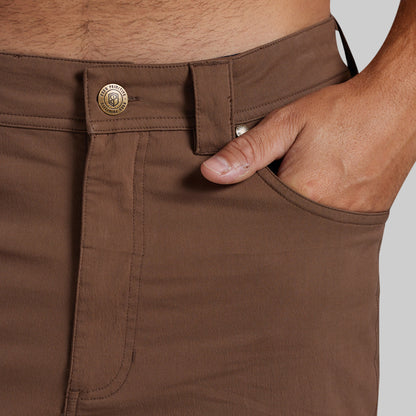 Close-up of a person wearing brown pants, showing the waistband, belt loop, front button, pocket, and hand in the pocket against a light gray background.