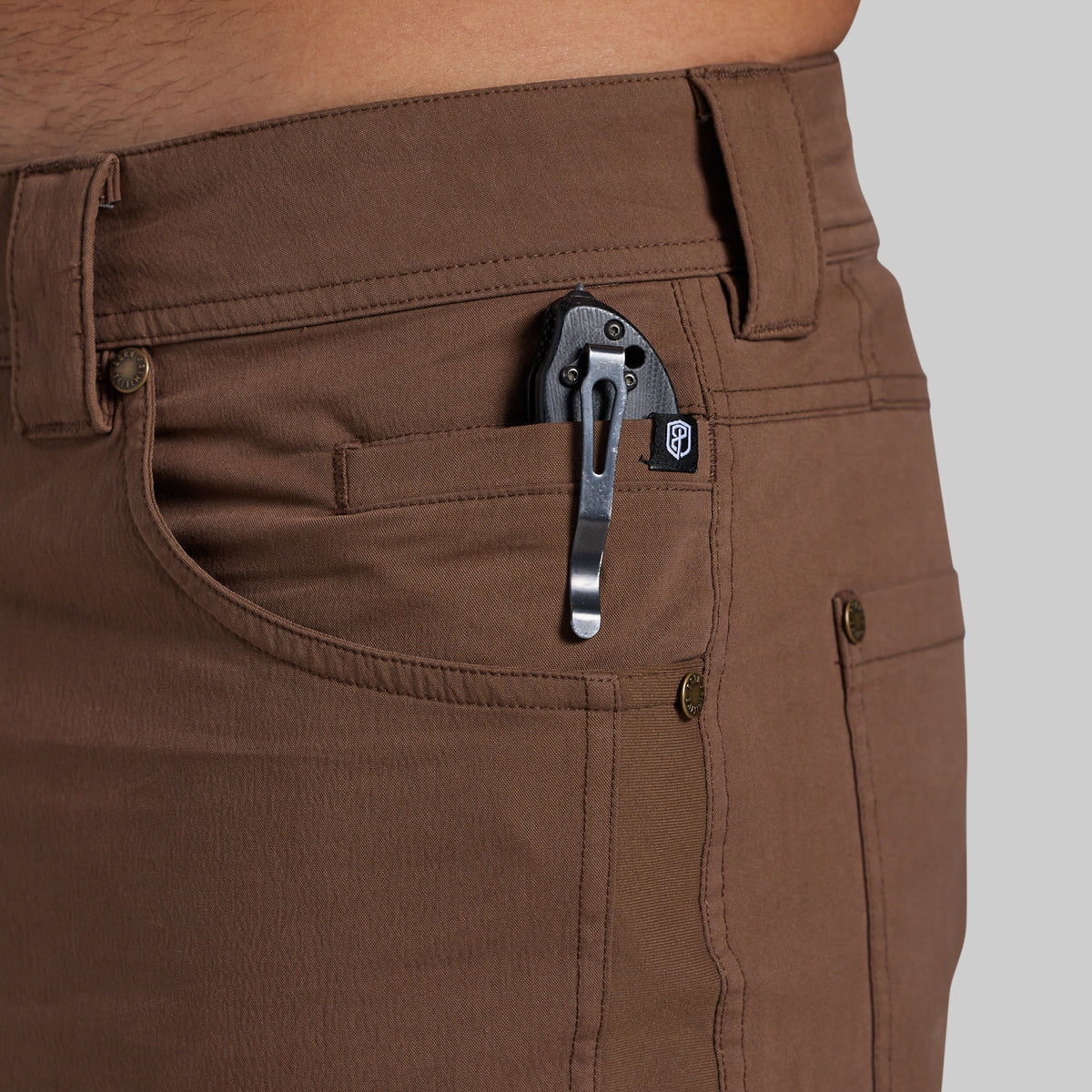 Close-up of a person wearing brown pants, showing the back pocket with a black metal pocket clip holding a small black accessory.