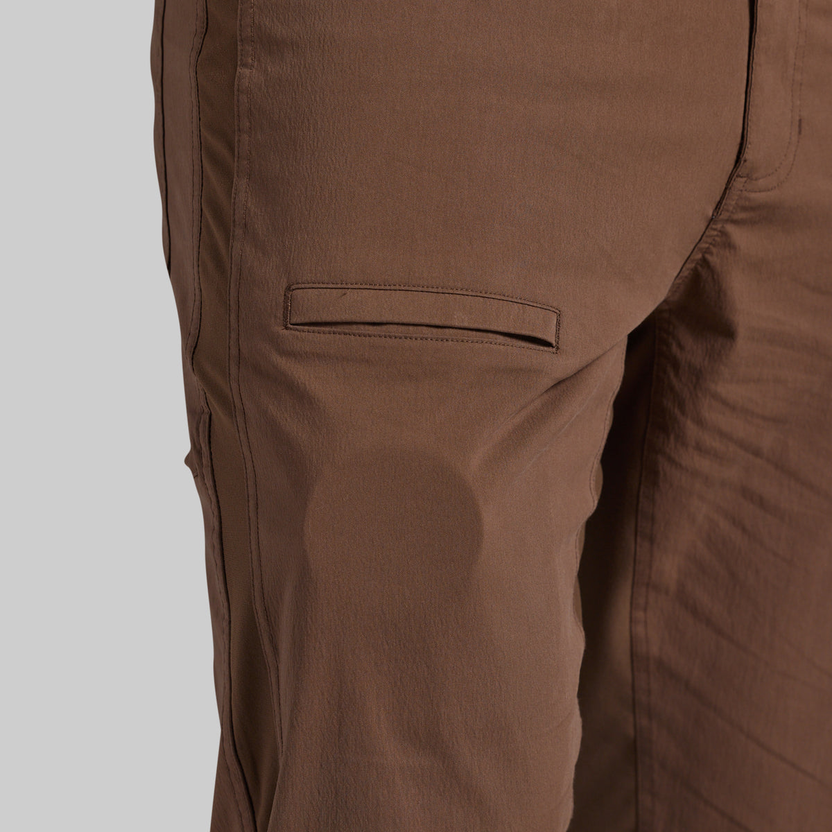 Close-up of brown pants showing a side pocket and fabric texture against a light gray background.