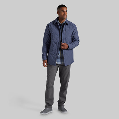 A man modeling a navy blue quilted jacket over a blue and gray plaid shirt, paired with gray pants and gray slip-on shoes, against a solid gray background.