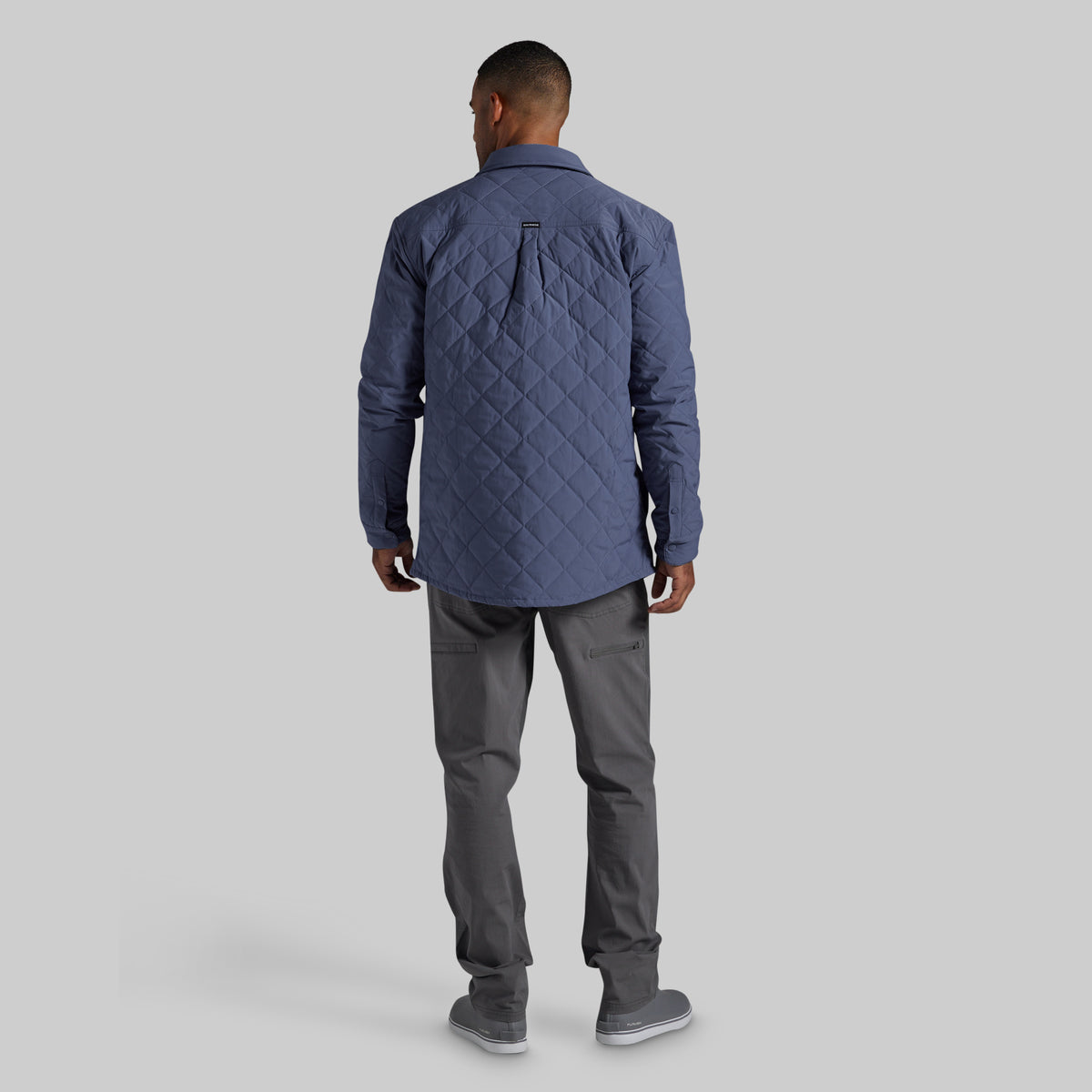 A man viewed from behind wearing a navy blue diamond-quilted jacket, dark gray trousers, and gray sneakers, standing against a solid light gray background.