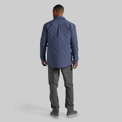A man viewed from behind wearing a navy blue diamond-quilted jacket, dark gray trousers, and gray sneakers, standing against a solid light gray background.